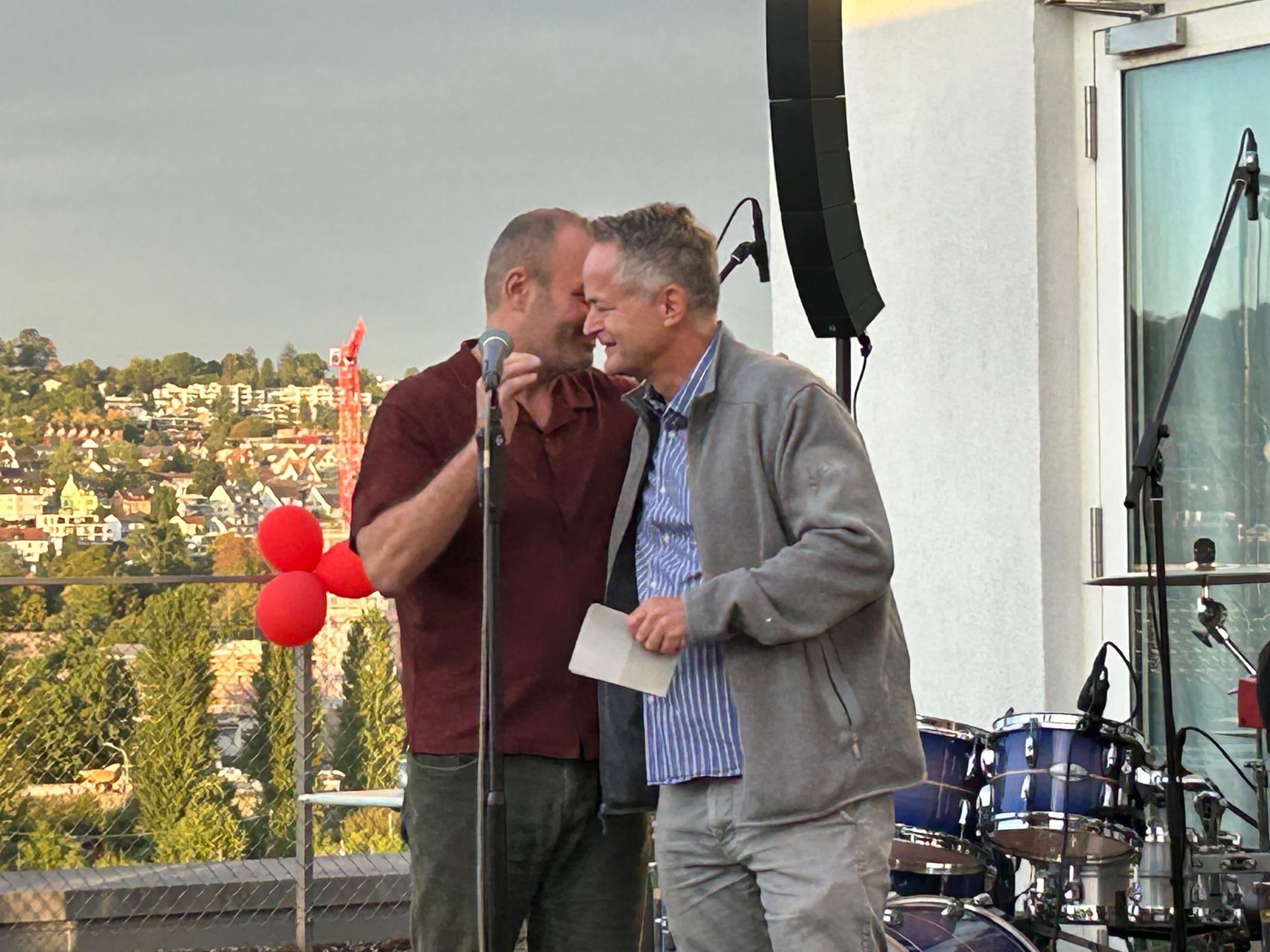 25 Jahre Kampagnenforum: Zwei Männer umarmen sich am Mikrofon auf der Terrasse